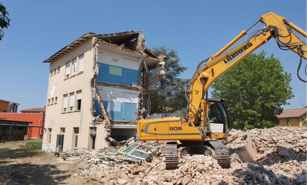 Escavatore giallo demolisce edificio, detriti e macerie intorno, cielo sereno.