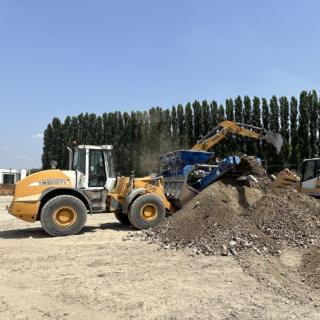 Ruspa sposta terra in un cantiere, alberi sullo sfondo, cielo sereno.