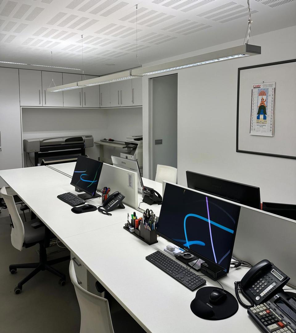 Ufficio moderno con scrivanie ordinate, computer, stampante e illuminazione a soffitto.